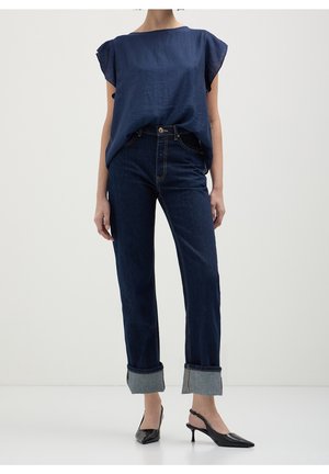 Femme portant un haut bleu marine à manches courtes, un jean foncé retroussé et des chaussures slingback noires à petit talon, debout devant un fond uni.