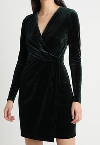 Mørkegrøn fløjlskjole med lange ærmer, v-udskæring og indpakningsdesign. Har en tætsiddende silhuet og glat tekstur.