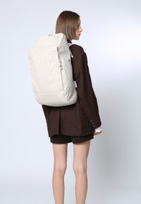 Weißer Rucksack mit Zugverschluss und Fronttasche, aus glattem Material gefertigt, mit dezenten runden Akzenten und verstellbaren Trägern.