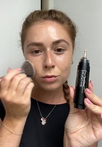 MAC Studio Fix foundation in a black container, held by a person applying product with a brush. Skin has a smooth, natural texture with freckles.