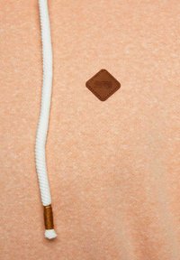 Pfirsichfarbener Sweater aus strukturiertem Stoff, mit einer weißen Kordel mit hölzernem Akzent und einem Logo-Patch aus braunem Leder.