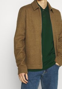 Homme portant une veste zippée marron sur un polo vert foncé et un jean bleu, se tenant avec une main dans la poche contre un fond blanc.