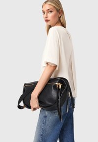 Zwarte leren handtas met gouden ritsen, rechthoekige vorm en lange bandjes. Model draagt een lichtbeige top met kanten rand en een blauwe spijkerbroek.