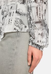 Durchsichtige Bluse mit monochromen abstrakten Zeichnungen, elastischen Manschetten und einem leicht gerafften Saum. Kombiniert mit hellgrauen Jeans.