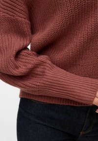 Adīta rozā krāsas džemperis ar teksturētu rakstu, ribveida manžetēm un brīva piegriezuma, kombinēts ar tumšām džinsa biksēm.