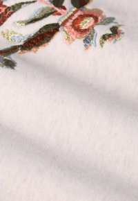 Tissu brodé avec des motifs floraux en rose, vert et or sur un fond clair, mettant en valeur une texture douce et des détails délicats.