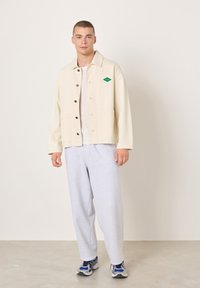 Beige Baumwolljacke mit Knopfleiste, zwei großen Taschen und grünem Diamantlogo. Hellgraue Jogginghose und blaue Sportschuhe getragen.