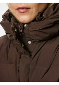 Brūna piedurkņu jaka ar augstu apkakli, ar rāvējslēdzēju un pogām ar snap tipa aizdari. Tam ir gluda tekstūra un smalkas šuvju detaļas.