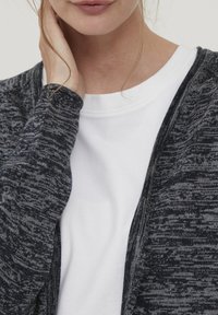 Cardigan gris foncé à motif avec un design en tricot texturé, superposé sur un t-shirt blanc uni. Gros plan sur l'encolure et la manche.