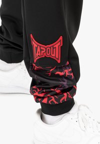 Crne trenirke s crvenim logom "TAPOUT" na bedru, ukrašene živopisnim crvenim i ljubičastim plamenovima na donjem dijelu noge. U paru s bijelim tenisicama.