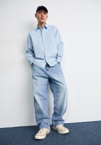 Lichtblauwe gestreepte overhemd met knopen, losse lichtblauwe denim jeans en beige sneakers met rode accenten. Model staat tegen een witte muur.