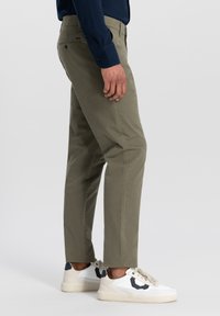 Olivinės spalvos chinos kelnės su slim fit modeliu, turinčios priekinės ir galinės kišenes, derinamos su baltomis sportbačiais su tamsiai mėlynais akcentais.