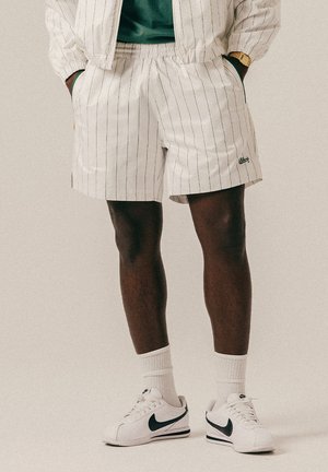 Osoba w białych spodenkach w cienkie paski, białych skarpetkach i białych butach Nike z czarnym logo, z rękami w kieszeniach, stojąca na jednolitym tle.