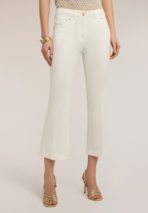 Vrouw draagt witte, hooggetailleerde, wijd uitlopende broek die net boven de enkel is afgeknipt, gecombineerd met gouden sandalen met hoge hak en gouden armbanden.