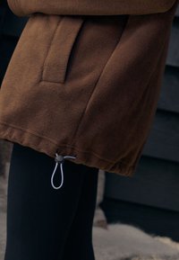 Brun fleecejakke med snøre i taljen med lukning og sidelomme, kombineret med sorte leggings.