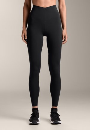 Magas derekú fekete leggings rugalmas anyagból. Simább textúrával és testhezálló fazonnal rendelkezik, fekete sportcipővel párosítva.