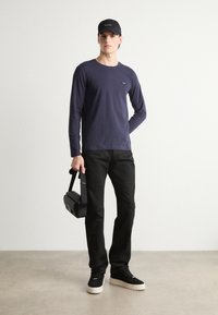 Μακριμάνικο ναυτικό πουκάμισο, μαύρο παντελόνι, μαύρα αθλητικά παπούτσια με λευκές σόλες και μαύρη τσάντα. Μινιμαλιστικός σχεδιασμός με στενή εφαρμογή και καλοκαιρινό στυλ.