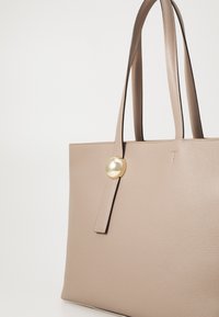 Sac fourre-tout en cuir beige avec une surface texturée, deux longues poignées et un accent circulaire doré à l'avant. Design simple et moderne.