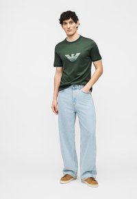 Νεαρός άνδρας στέκεται με το ένα χέρι στην τσέπη φορώντας σκούρο πράσινο t-shirt με λογότυπο, ανοιχτό μπλε τζιν παντελόνι με φαρδιά μπατζάκια και καφέ αθλητικά σε μονόχρωμο φόντο.