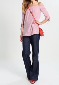 Donna che indossa un top rosa a righe scollato, jeans larghi scuri, borsa a tracolla rossa e sandali aperti rossi, in piedi con una mano sui fianchi.