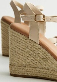 Sandale compensée beige avec une semelle en jute tressé, des brides en cuir et un semelle intérieure ronde rembourrée avec un subtil accent de boucle dorée.