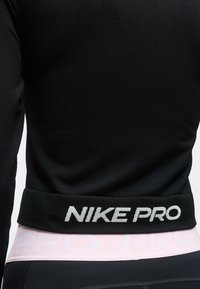 Μαύρο αθλητικό τοπ Nike Pro φορεμένο πάνω από ροζ ζώνη μέσης με μερικώς ορατό λευκό κείμενο "PRO" στα αθλητικά κολάν.