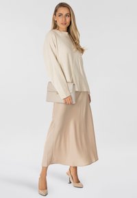 Maglione beige lavorato a maglia con orlo a coste, abbinato a una gonna beige fluida di lunghezza al ginocchio. Una borsa grigia con una chiusura accentata in oro è appoggiata di lato.