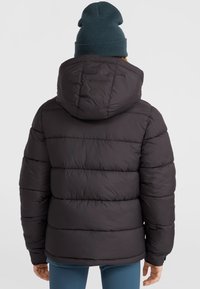Svart puffjacka med horisontell quiltad design och huva, bärs med en mörkblå mössa, sedd från baksidan. Slät textur.