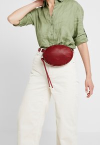 Borsa a tracolla ovale in pelle rossa con zip, patta appesa e logo stampato, indossata sopra una camicia in lino verde e pantaloni in corduroy beige.