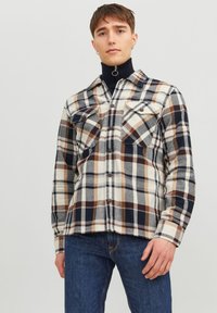 Chemise en flanelle avec un motif à carreaux multicolore dans des tons de crème, marron, marine et gris. Elle possède deux poches poitrine et un col pointu.