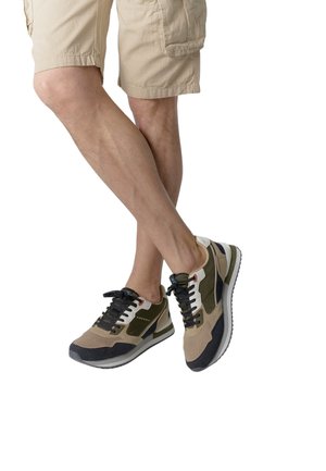 Man draagt beige korte broek en multicolor sneakers met zwarte veters, benen gekruist tegen een witte achtergrond.