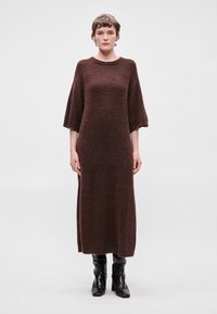 Brūns, garām piedurknēm adīts kleitas zīmolējums ar brīvu piegriezumu un apaļu kakla izmēru, ar teksturētu virsmu. Saskaņots ar melniem ceļgala augstiem zābakiem.