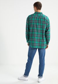 Camicia a quadri verde con maniche lunghe, dotata di tasche e polsini con bottoni, abbinata a jeans blu e sneaker bianche.