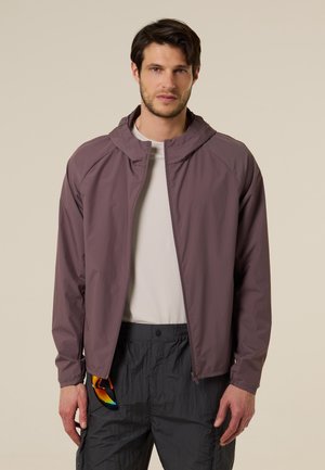 Man draagt een mauve ritssluitingvest over een licht shirt, zwarte broek, houdt reflecterende zonnebril in linkerhand vast, staat voor een beige achtergrond.