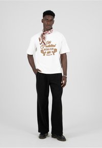 Valge ülisuur t-särk pruunide graafiliste trükkidega, koos lõdvate mustade pükstega ja mustriga salliga kaelas. Mustad kingad täiendavad välimust.