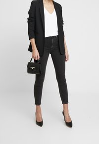 Černý blazer přes bílou tričko s výstřihem do V, spárovaný s černými skinny jeans a černými špičatými podpatky. Drží malou černou kabelku s embosovaným vzorem.