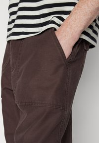 Pantalon marron foncé au toucher lisse, doté de poches latérales et de surpiqûres, associé à une chemise rayée noire et blanche.