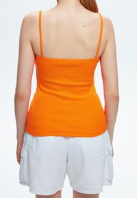 Débardeur orange côtelé avec des bretelles fines, vu de l'arrière, associé à un short cargo blanc avec des poches latérales.