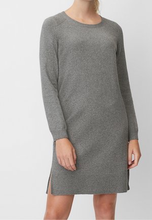 Woman wearing a long-sleeve, knee-length grey knit dress with side slits, standing against a plain light background.