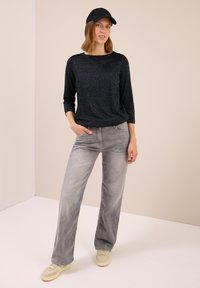 Schwarzes, glitzerndes Langarmoberteil, kombiniert mit hellgrauen, weiten Jeans und cremigen Sneakers. Das Modell trägt eine schwarze Mütze und steht vor einem neutralen Hintergrund.
