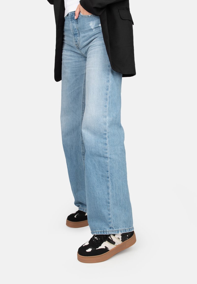 Jeans a gamba larga azzurri chiari con una finitura sbiadita e orlo sfilacciato, abbinati a sneakers bianche e nere con superficie testurizzata.