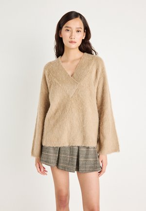 Jeune femme portant un pull en V beige et moelleux, accompagné d'une jupe plissée écossaise courte, se tenant devant un fond blanc uni.