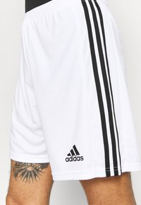 Pantaloni sportivi bianchi realizzati in tessuto leggero, con tre strisce nere sui lati e un logo adidas sull'orlo inferiore.