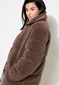 Cappotto in pile marrone con un design gonfio, colletto alto e tessuto testurizzato. Presenta un colletto liscio e in contrasto di una tonalità più scura.