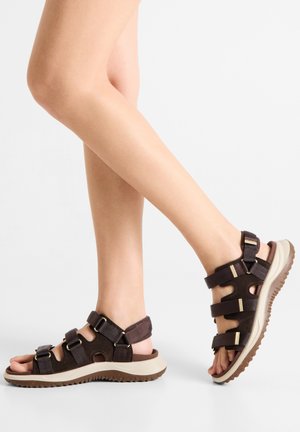 Gambe con sandali marroni con punta aperta, tre cinturini regolabili e una suola spessa bianca e marrone, in piedi su un pavimento bianco.