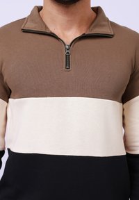 Pullover avec un col zippé, composé de trois blocs de couleur : marron, crème et noir. Fabriqué en tissu doux avec une finition texturée.