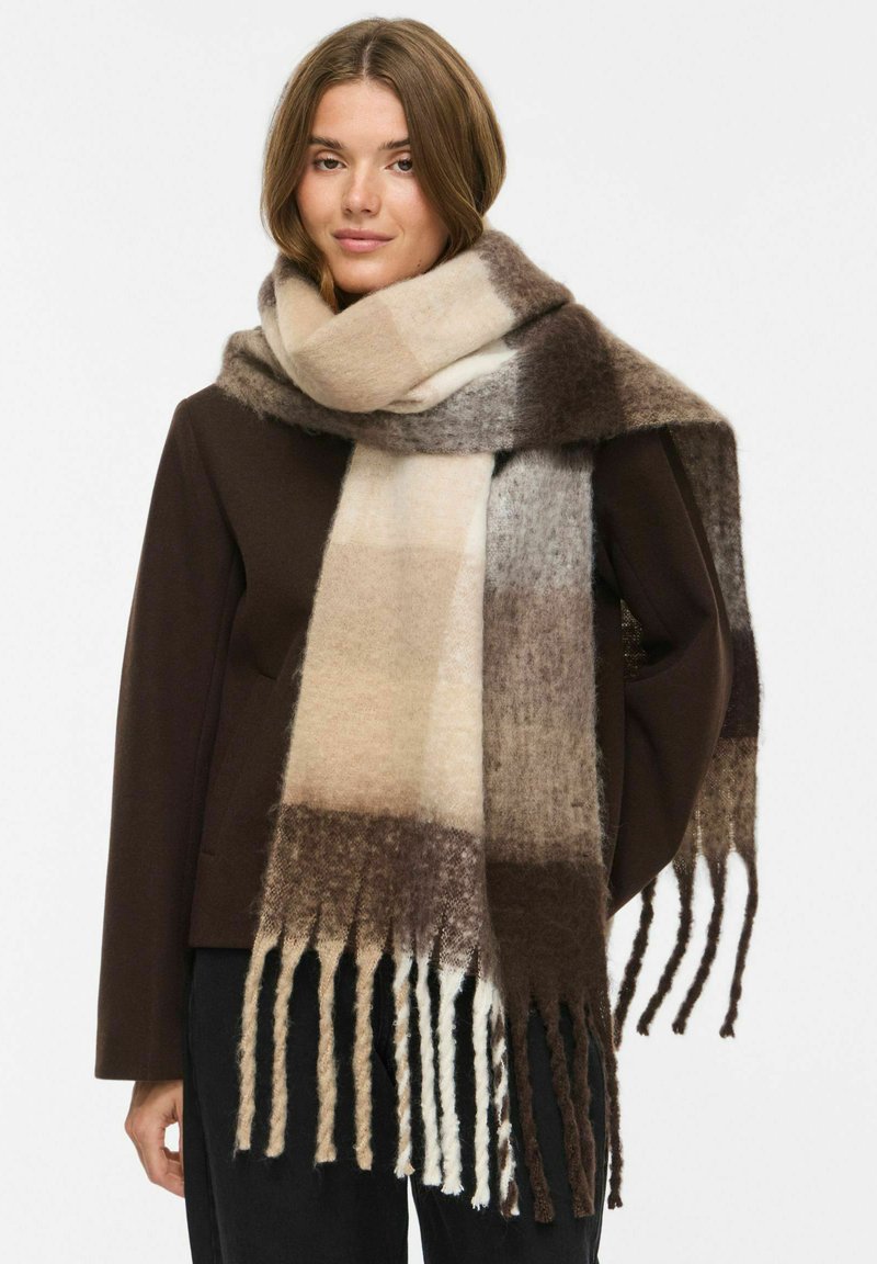 Femme portant un manteau marron foncé et une grande écharpe douce à motif blocs beige, gris et marron avec de longues franges.