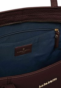Interior de un bolso de mano de cuero marrón oscuro con forro de tela azul marino, un bolsillo con cremallera y una pequeña etiqueta de piel de la marca en el interior.
