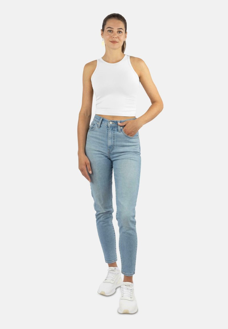 Woman standing wearing a white sleeveless crop top, light blue high-waisted jeans, white sneakers, and yellow earrings against a white background.