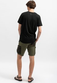 Zwarte katoenen t-shirt met een ronde hals, gecombineerd met olijfgroene cargoshorts met meerdere zakken en zwarte accenten. Platte sandalen zijn zichtbaar.